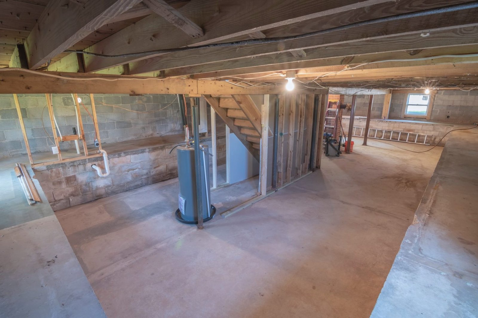 An unfinished basement with exposed joists, block walls and a blue water heater standing by the stairs