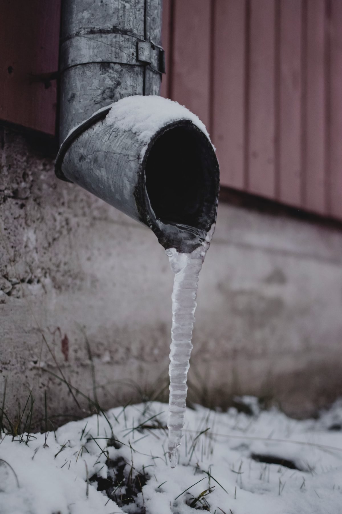 A metal downspout with a column of ice hanging from its mouth above snow