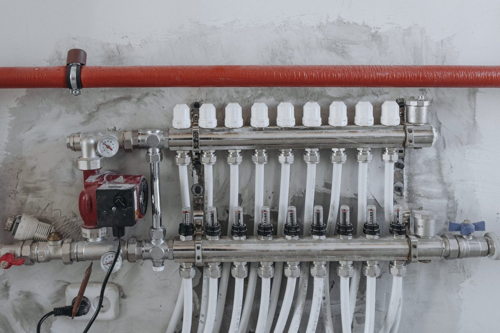 A wall-mounted heating manifold with a row of flow tubes, a red circulator pump and a thermometer