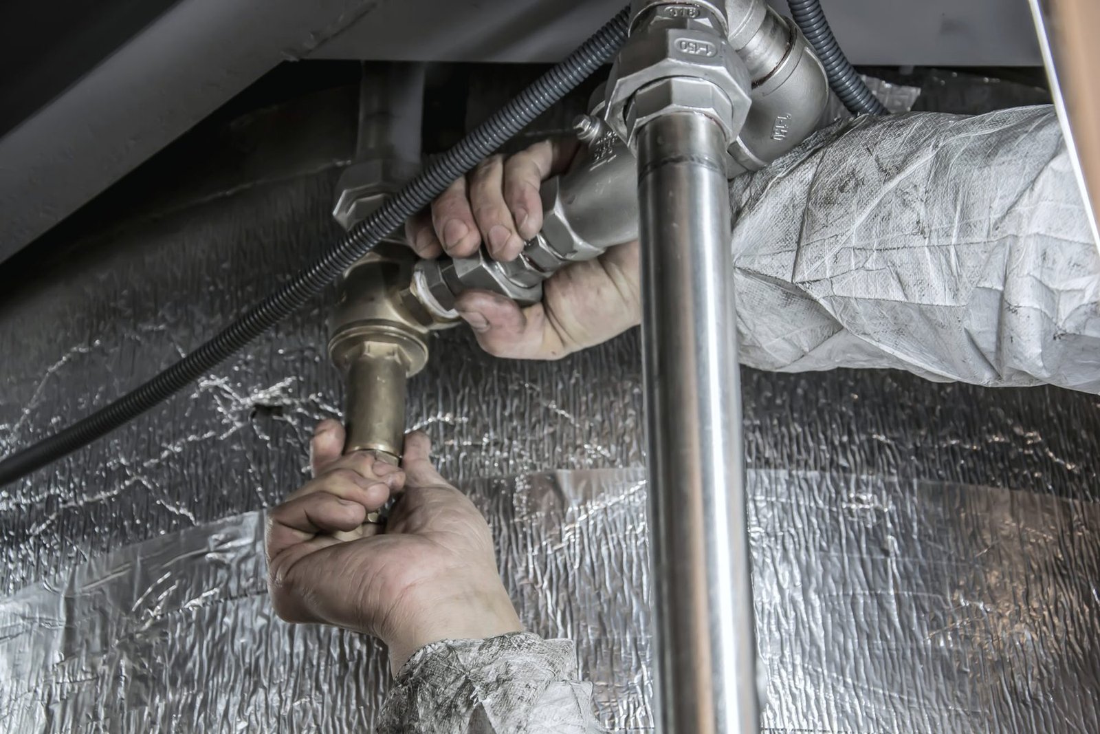 Bare hands tightening a threaded steel fitting on a pipe run beside foil-wrapped insulation