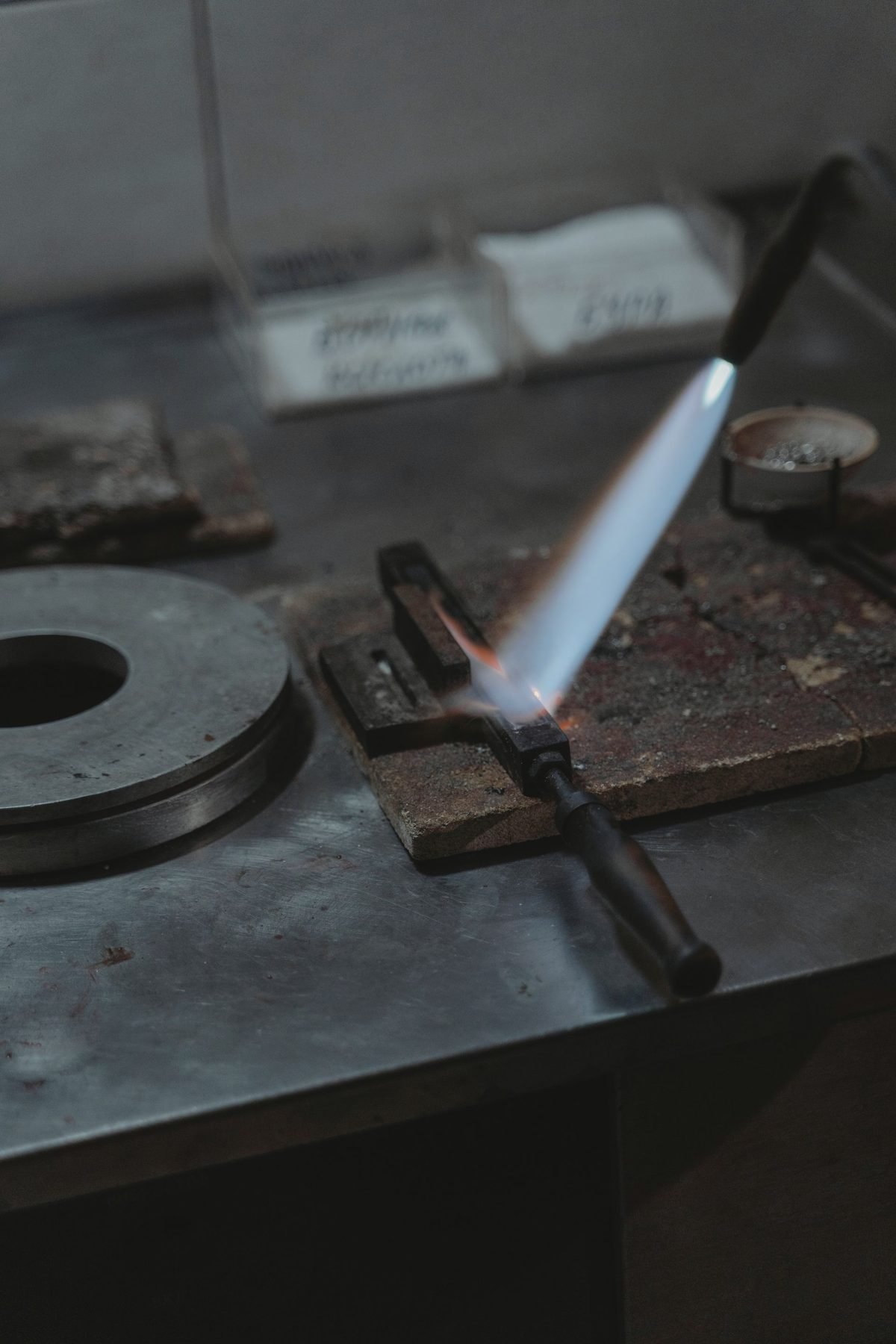 A lit gas torch with a narrow blue flame resting on a steel bench