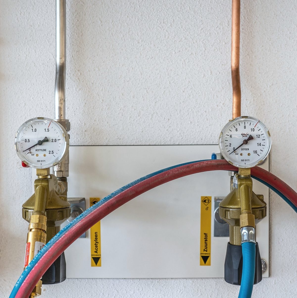 Two brass gas regulators with gauges and red and blue hoses mounted on a white wall