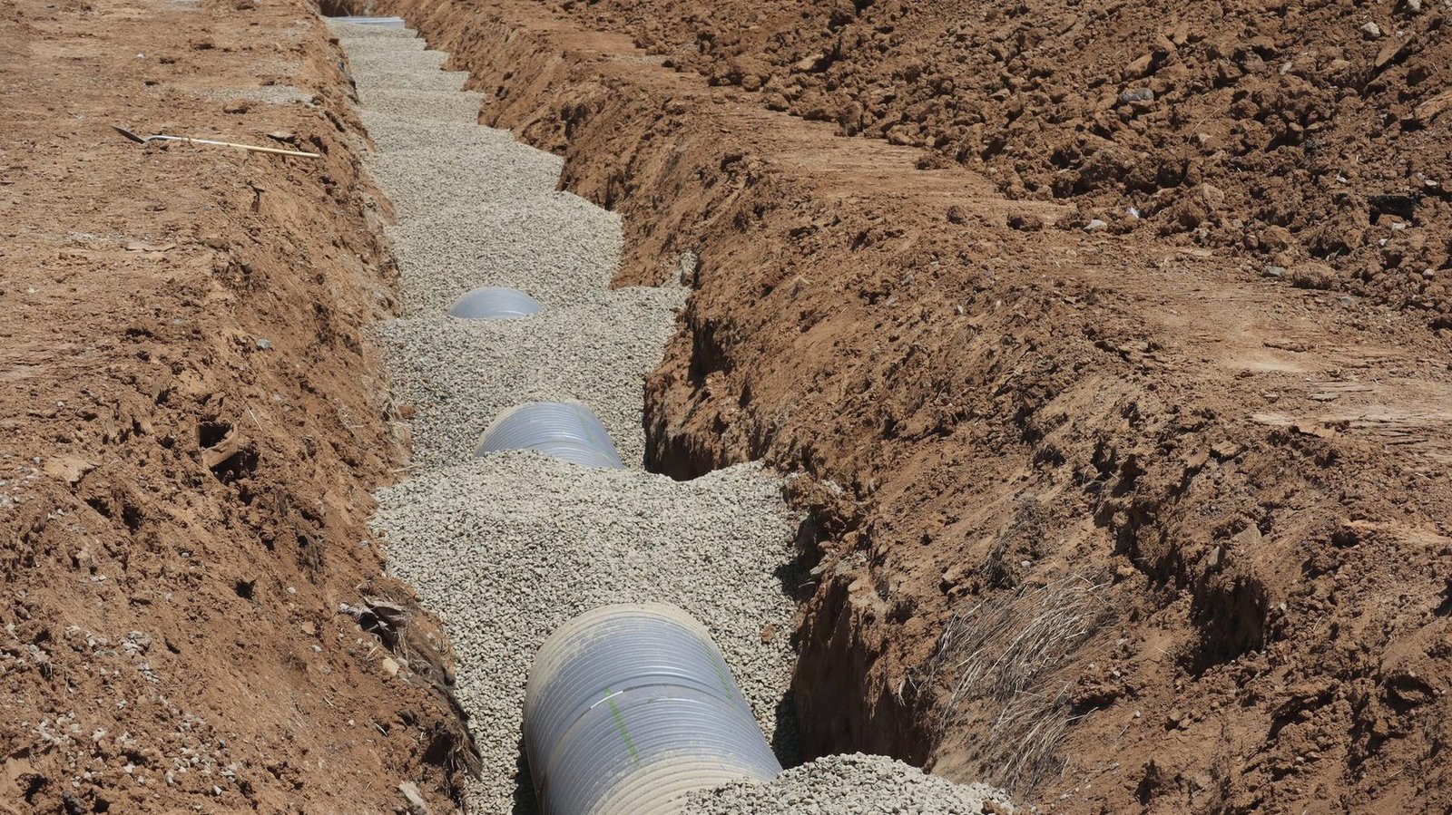 A long open trench in brown soil with grey drain pipe laid on a gravel bed